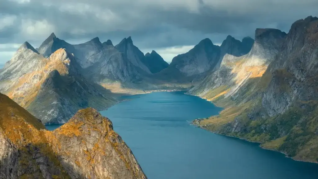 Aerial view of Lofoten Islands in Norway showcasing jagged peaks, mountains, and a fjord snaking through under a sunlit autumn sky, ideal for Scandinavian tourism.