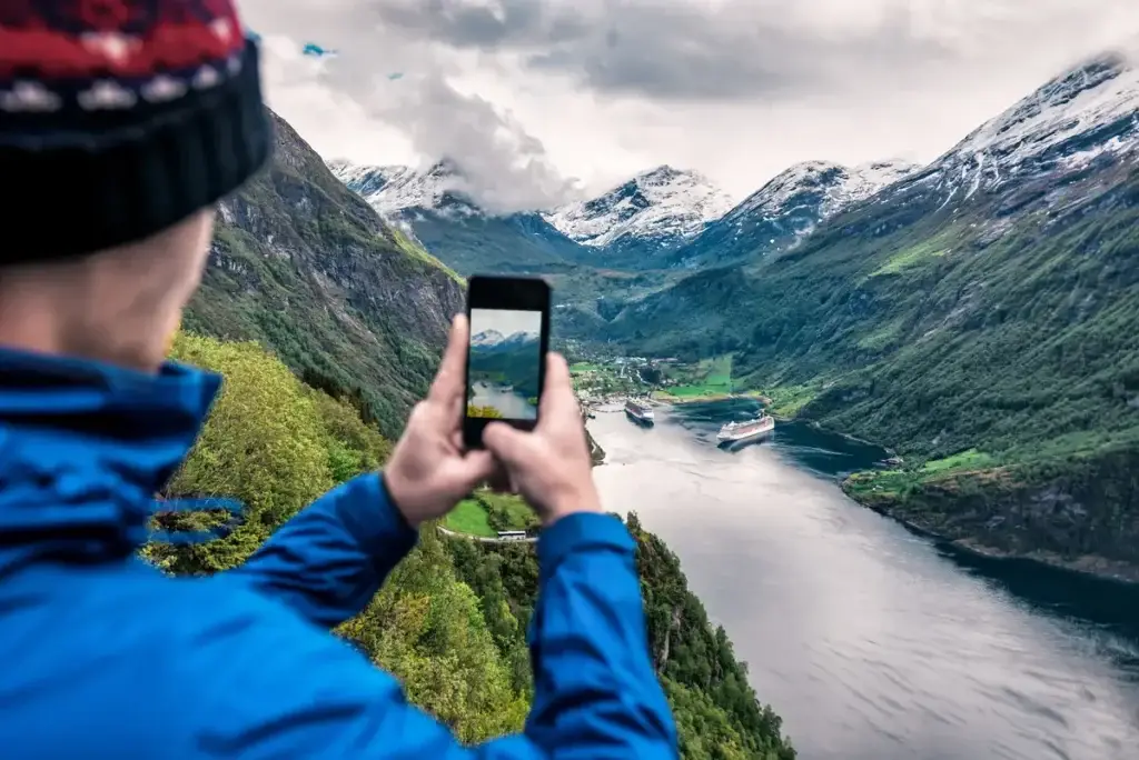 Tourist captures breathtaking view of Geiranger Fjord in Norway using smartphone, showcasing snow-capped mountains and serene waters with cruise ships.