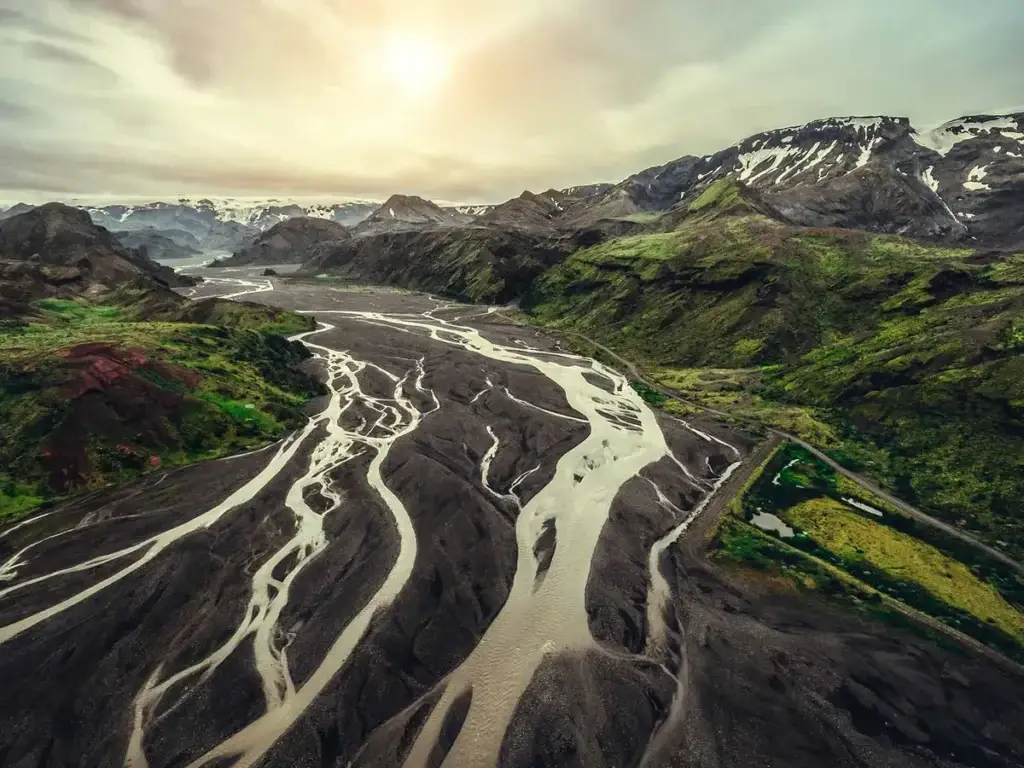Aerial view of a winding river flowing through a dramatic Icelandic landscape with snow-capped mountains and lush greenery under a bright sun, from Scandinavia Private Tours' 16-Day Iceland Tour.