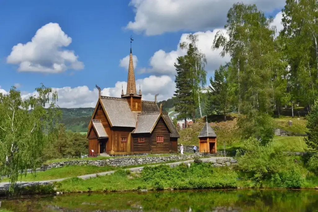 Traditional wooden stave church in Skanzen, Lillehammer, Norway, surrounded by lush greenery and mirrored in a tranquil pond under a blue sky with clouds.
