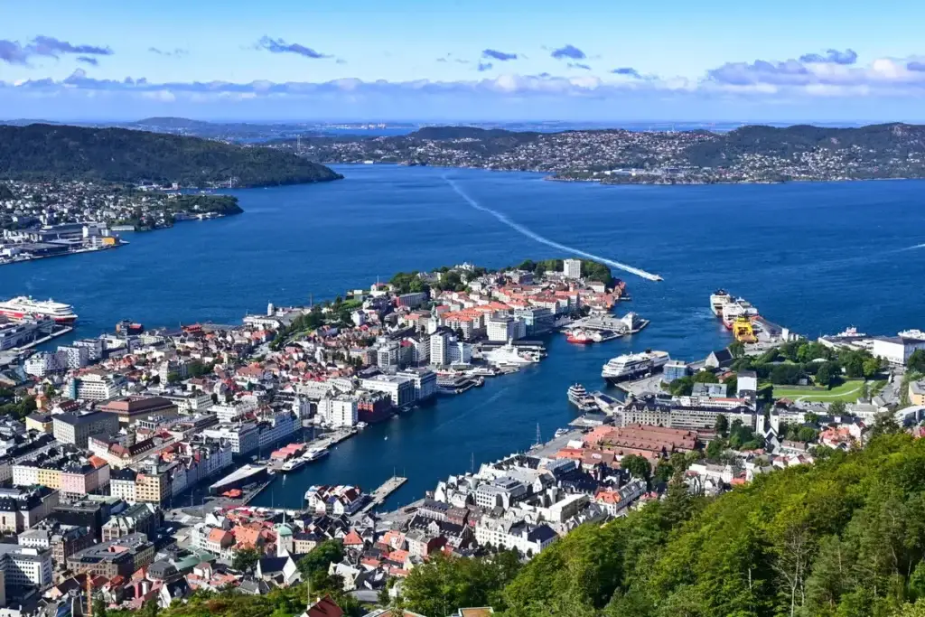 Aerial view of Bergen city and harbour in Norway, showcasing the dense architectural layout along the waterfront with multiple ships, including a ferry entering the port, surrounded by lush green hills under a clear sky.