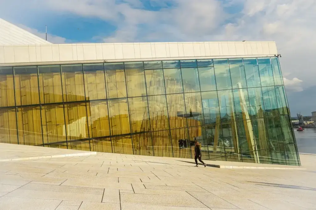 Tourist walking by the modern architecture of Oslo Opera House in Norway, reflecting cityscape in its glass facade.