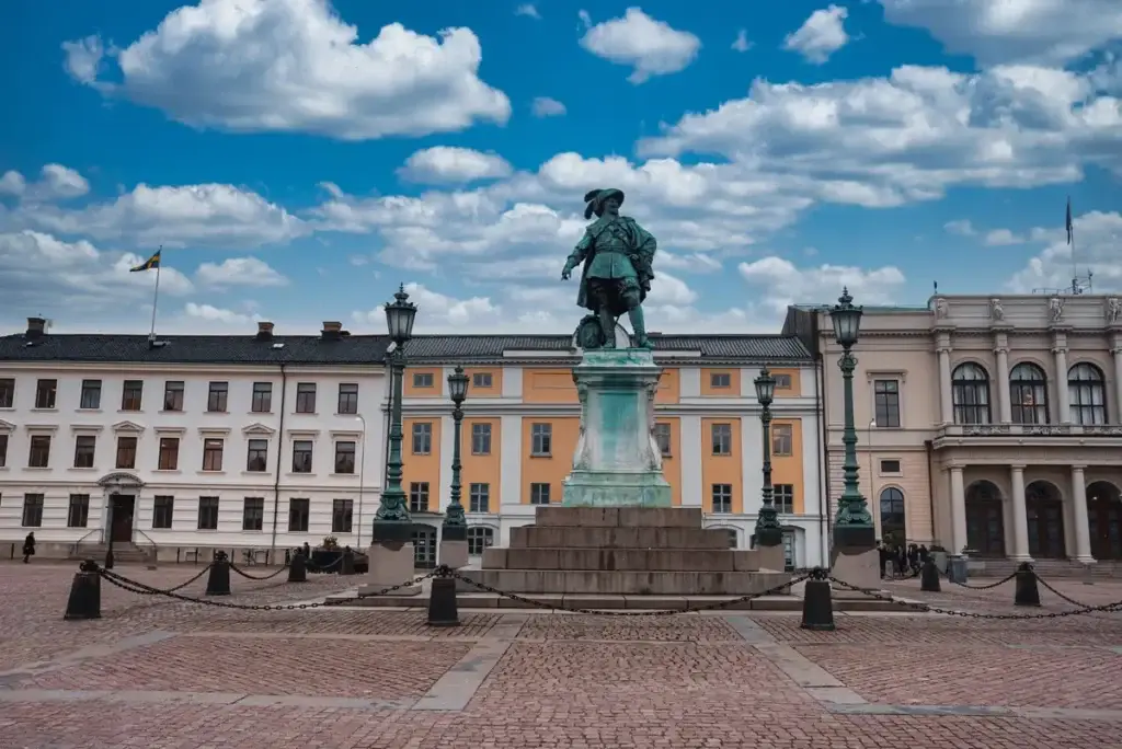 Bronze statue of a historical figure in Burunnsparken, city center of Gothenburg, Sweden, flanked by classical architecture under a blue sky with fluffy clouds.