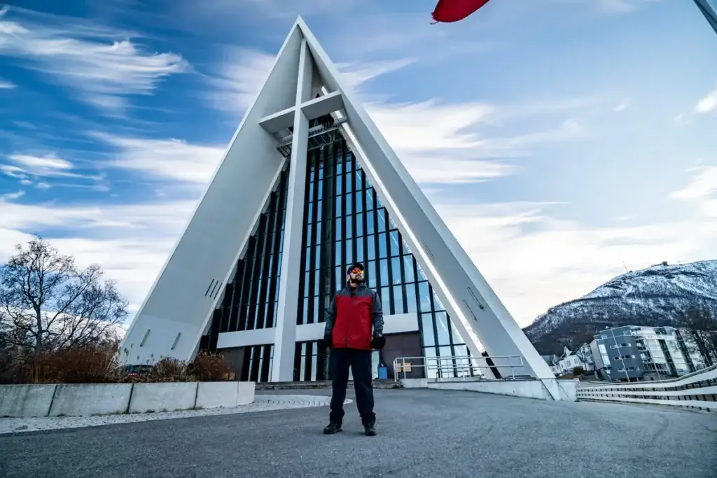Young man with beard in red jacket standing in front of the iconic Arctic Cathedral in Tromso, Norway, with scenic snowy mountains in the background.
