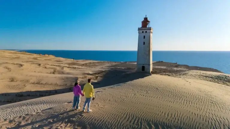 Couple walking toward iconic white lighthouse on sandy shores of Denmark under clear blue sky