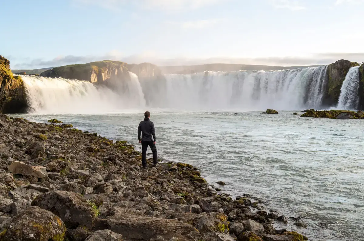 Man observing the majestic Godafoss waterfall in Iceland, surrounded by rugged terrain and cascading waters under a clear sky.