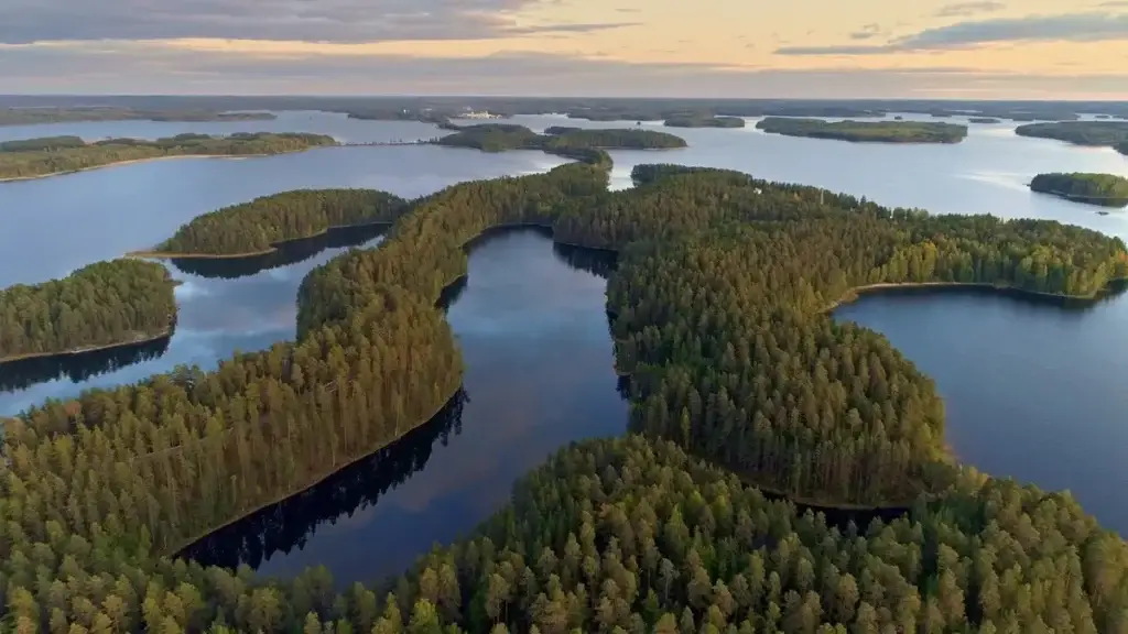 Aerial view of a serene Finnish lakeland at sunset, showcasing lush green islands and the tranquil reflection of clouds on the water, capturing the essence of Finland’s natural beauty in a classical landscape.