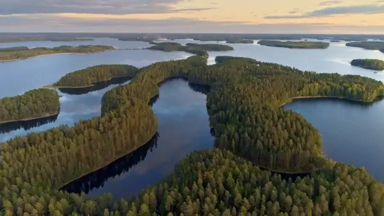 Aerial view of a serene Finnish lakeland at sunset, showcasing lush green islands and the tranquil reflection of clouds on the water, capturing the essence of Finland’s natural beauty in a classical landscape.