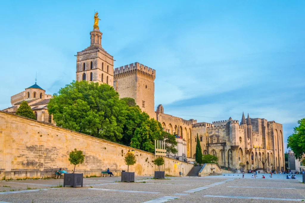 Sunset view of Palais des Papes and the cathedral in Avignon, France, featuring historic Gothic architecture and a statue atop a bell tower.