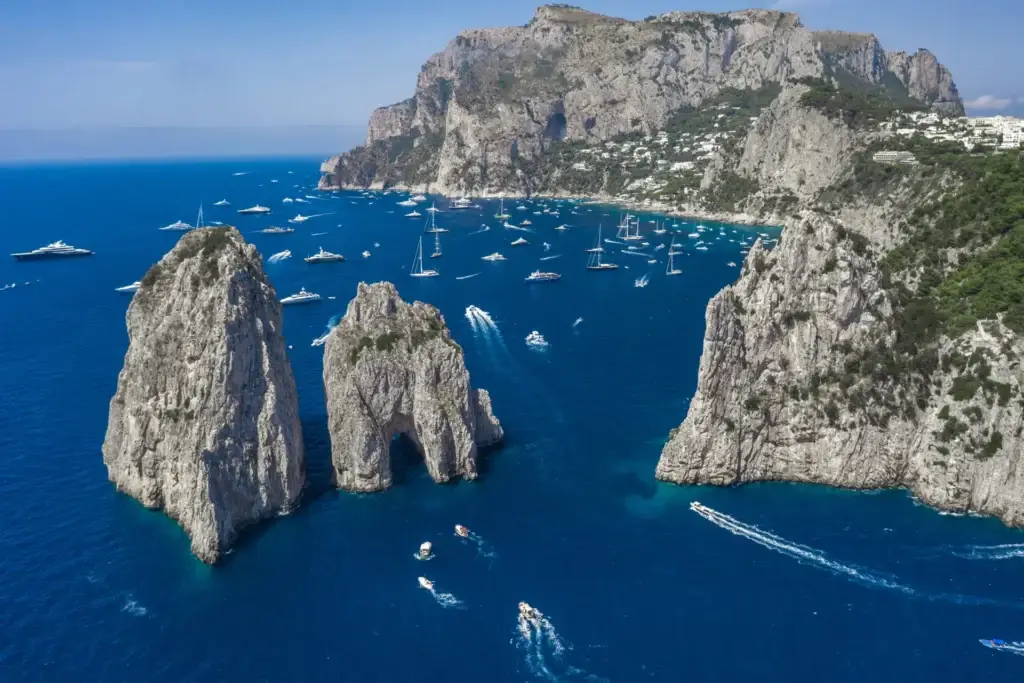 Aerial view of the dramatic Faraglioni rocks and luxury yachts sailing the clear blue Tyrrhenian Sea off Capri Island, a popular tourist destination in Italy.