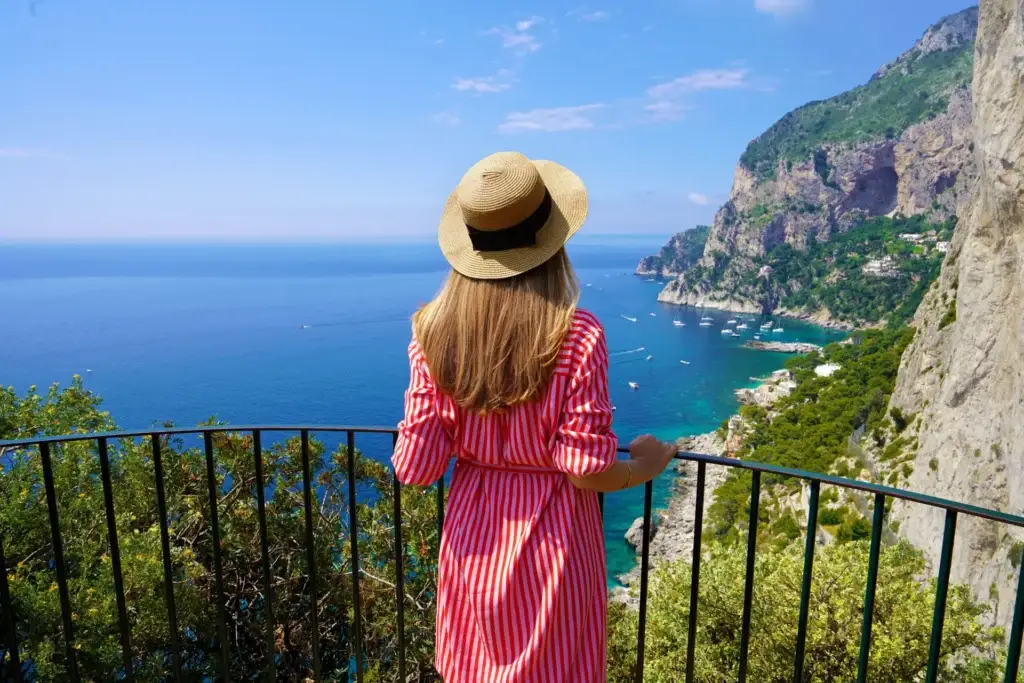 Young woman in a striped dress and sun hat admiring the panoramic view of the sea and cliffs from Capri Island, Italy.