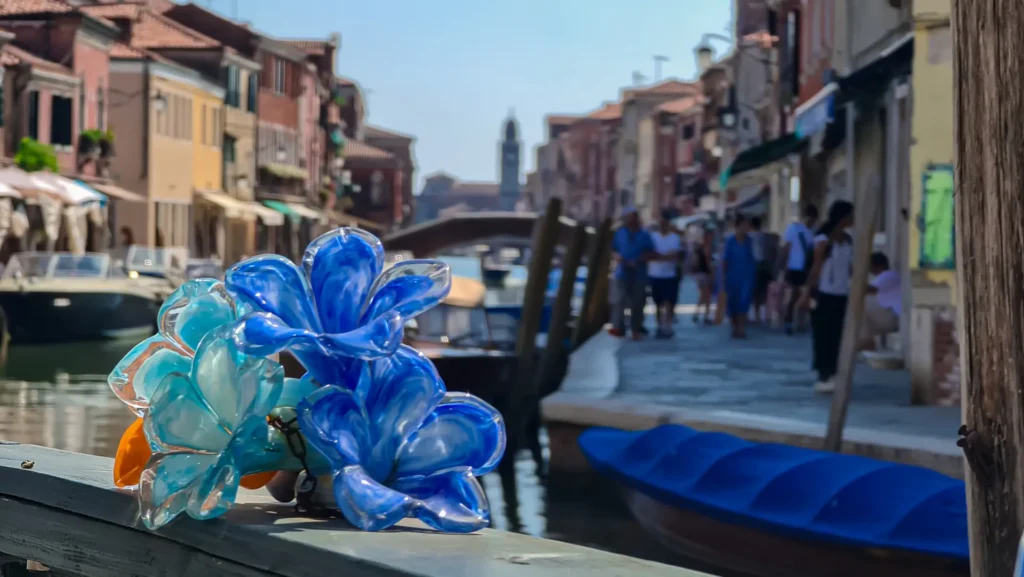 Colorful Murano glass flower in the foreground with a scenic backdrop of the clock tower and Ponte San Pietro Martire bridge over the canal Rio dei Vetrai on the island of Murano near Venice, Italy.