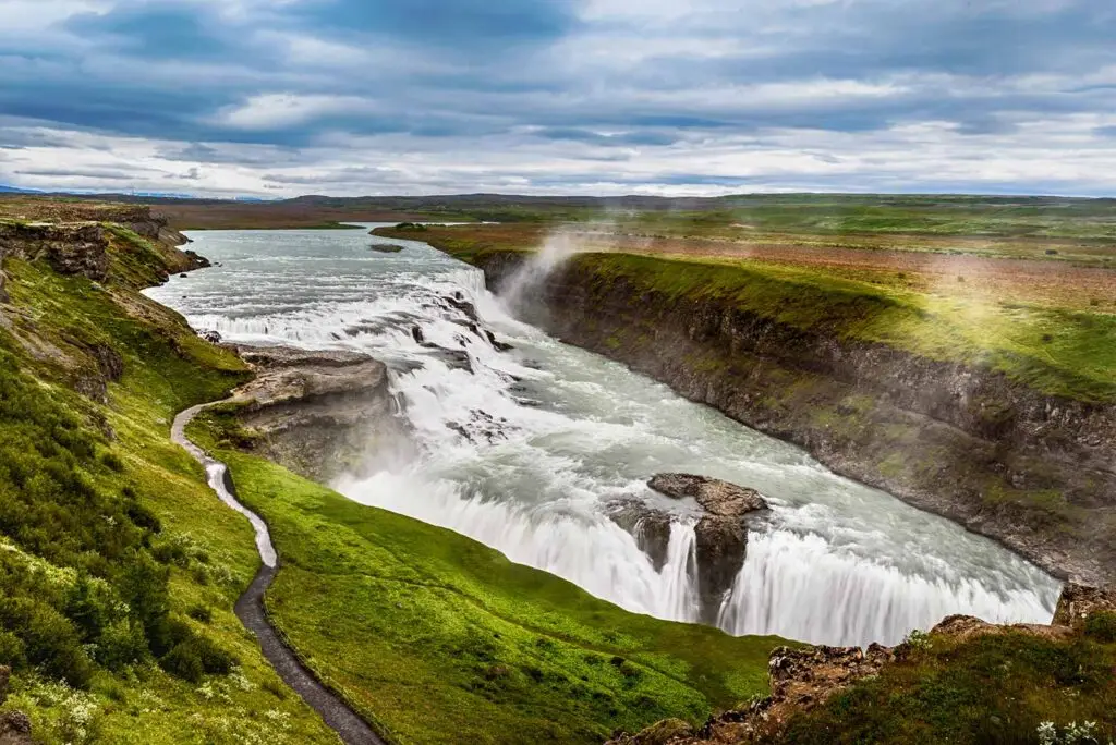 Scenic view of Gullfoss Waterfall along Iceland's Golden Circle Route, showcasing powerful cascades and lush green landscapes under a cloudy sky.
