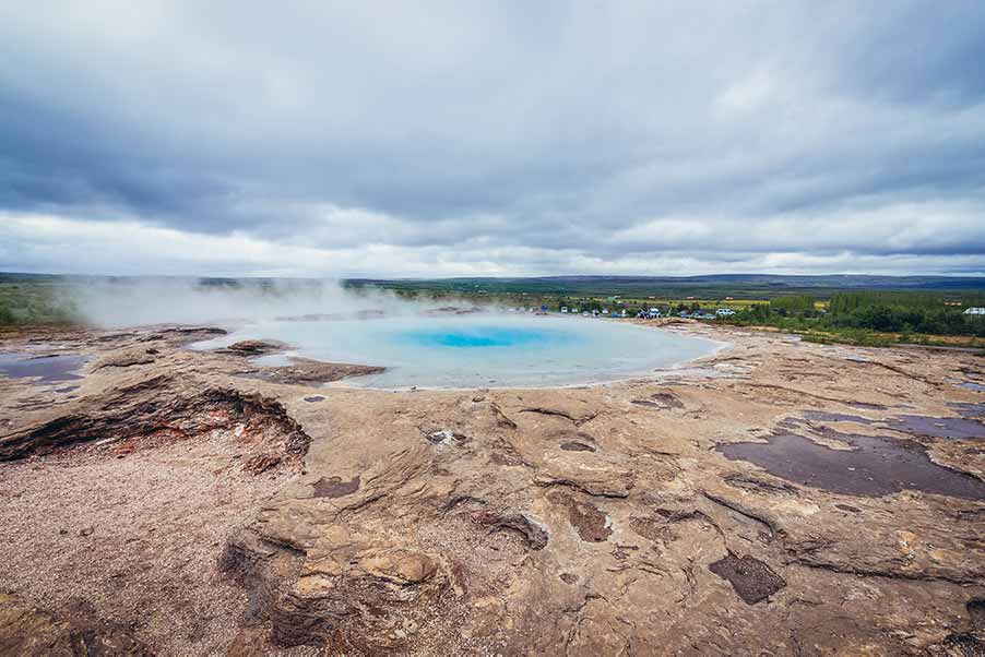 View of the Great Geysir in the geothermal area of Haukadalur Valley, Iceland, showcasing the vibrant blue water and steam rising against a dramatic sky.