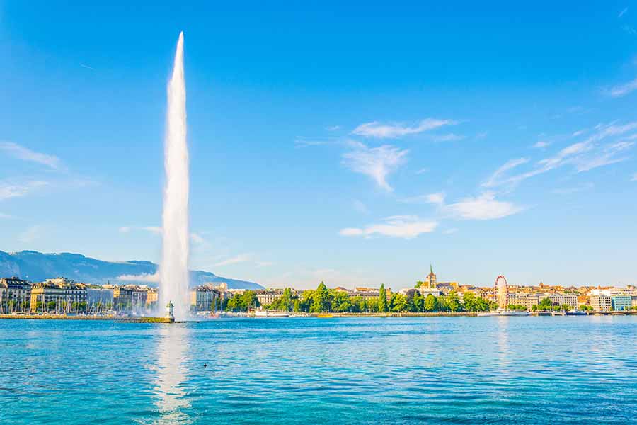 Iconic Jet d'Eau fountain spraying high into the blue sky on Lake Geneva with Geneva cityscape and mountains in the background, Switzerland.