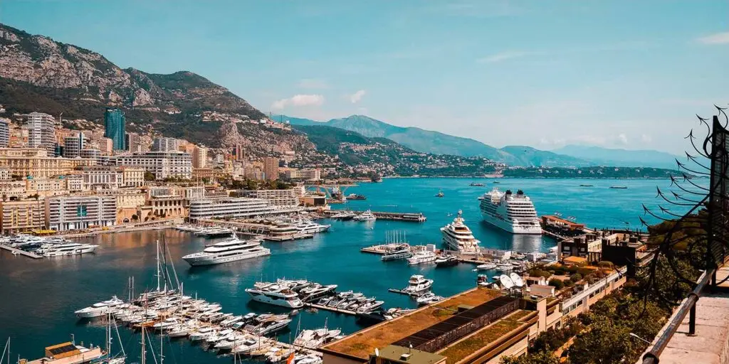 Panoramic view of Monte Carlo, Monaco, showing luxury yachts docked in the harbor with modern buildings and mountains in the background.