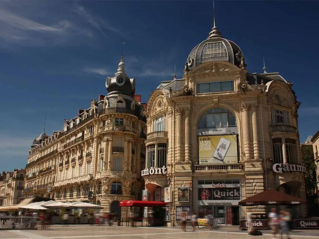 Historic architecture and bustling street life at Place de la Comedie in Montpellier, France, showcasing elegant Beaux-Arts buildings under a clear blue sky.