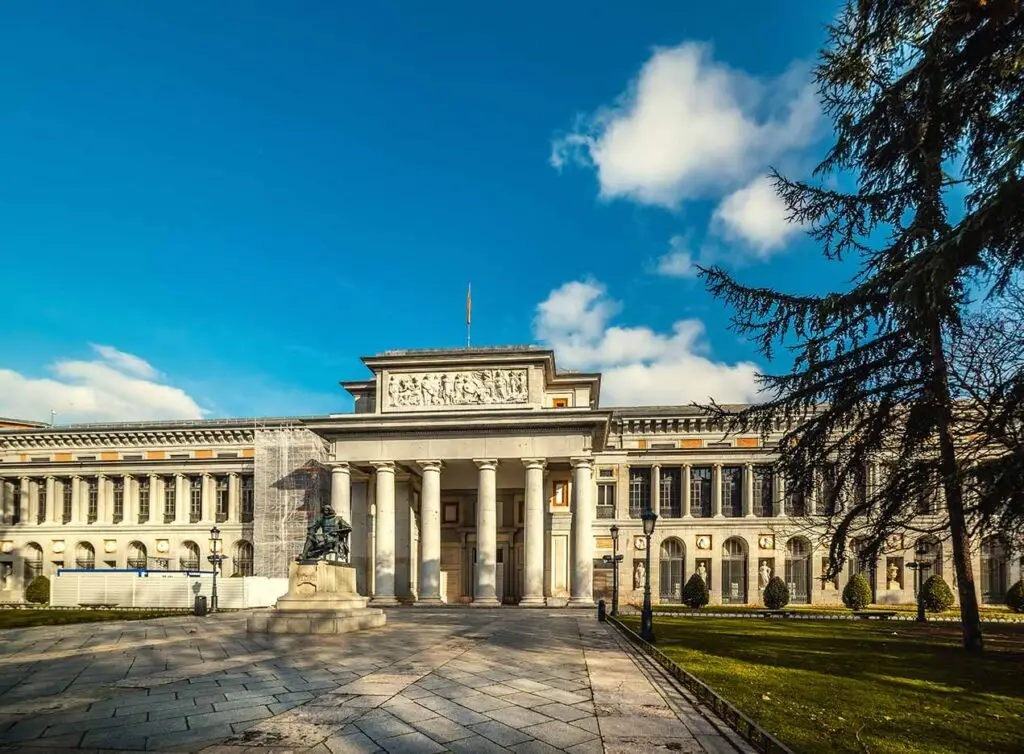 Exterior view of the Prado Museum in Spain showcasing classical architecture with columns and sculptures under a clear blue sky.