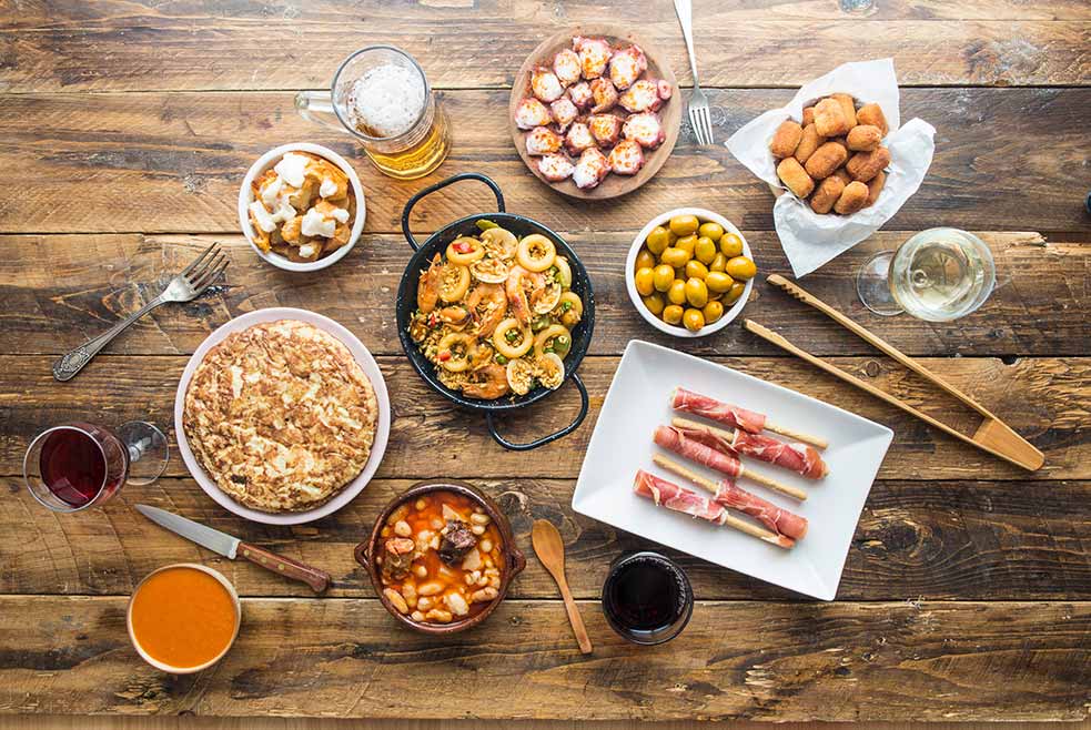 Traditional Spanish cuisine spread on a rustic wooden table, featuring dishes such as paella, tortilla Española, croquetas, and tapas, complemented by local wines and beer, perfect for gastronomic tourism in Spain