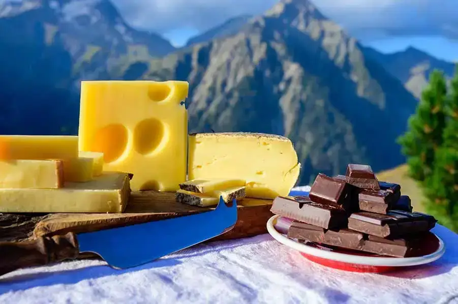 Assorted Swiss cheeses and chocolate bars on a wooden table with scenic Swiss mountains in the background.