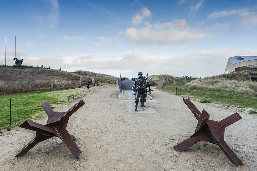 Historical memorial at Utah Beach depicting the D-Day invasion, with sculptures of soldiers and defensive obstacles, under a cloudy sky in Normandy, France.