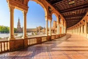 Scenic view of the iconic Plaza de Espana in Seville, Spain, showcasing the detailed architecture of arched bridges, ornate buildings, and vibrant ceramic tiles under a sunny sky.
