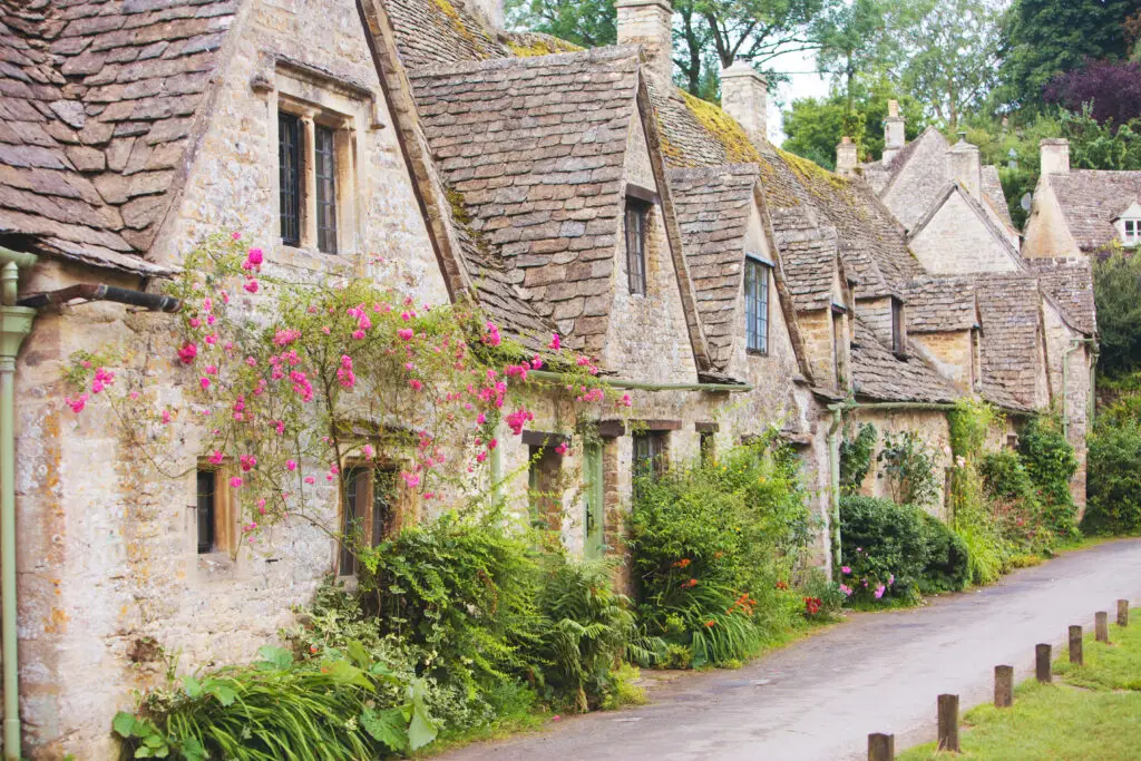 Historic stone cottages of Arlington Row in Bibury, Cotswolds, England surrounded by colorful flowers and greenery, a popular tourist attraction featured on Europe private tours
