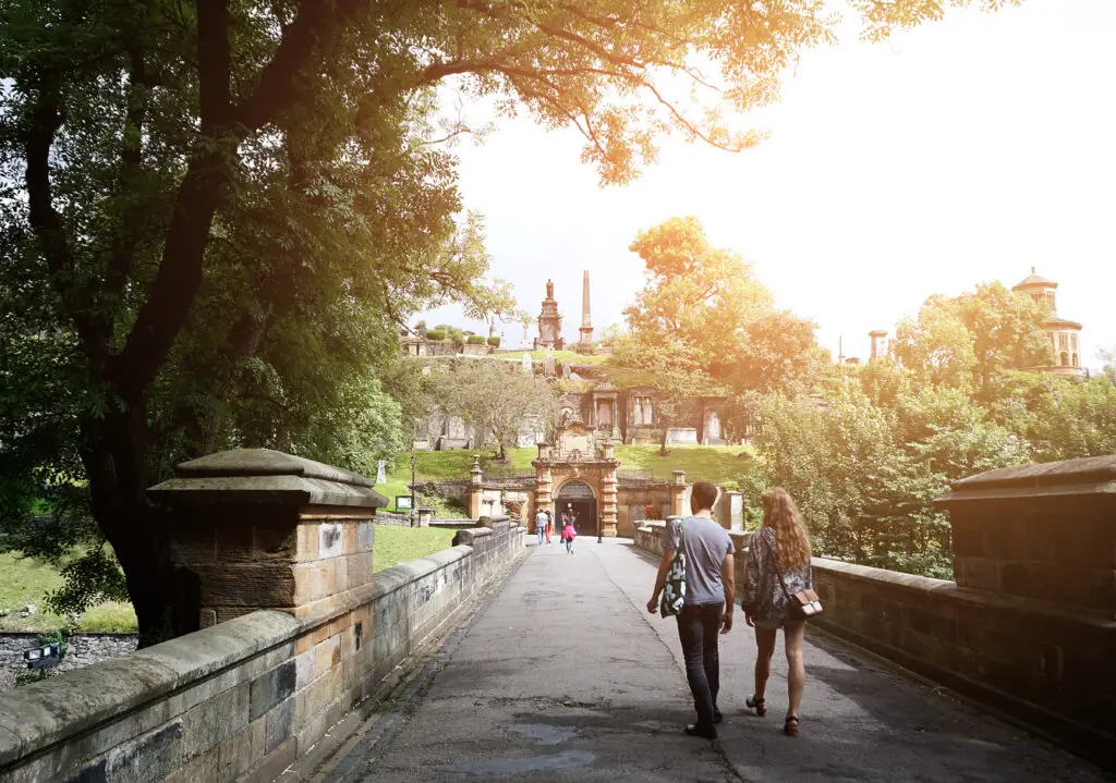 Tourists walking across a stone bridge toward the historic Glasgow Necropolis in Scotland surrounded by lush greenery and monuments at sunset