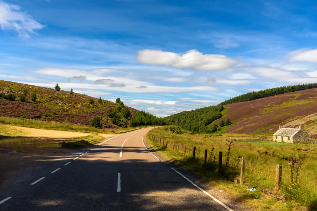 Scenic countryside road through Cairngorms National Park in Scotland with heather-covered hills, green forests, and a traditional stone cottage under a bright blue sky