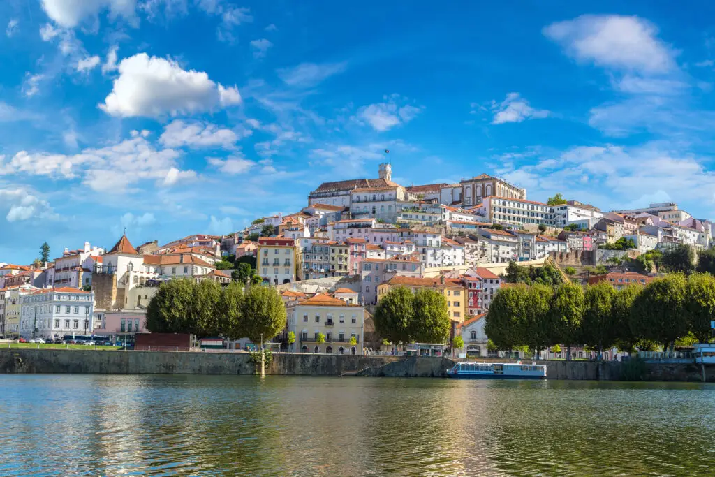 Scenic view of Coimbra, Portugal with historic buildings on a hillside overlooking the Mondego River under a bright blue sky