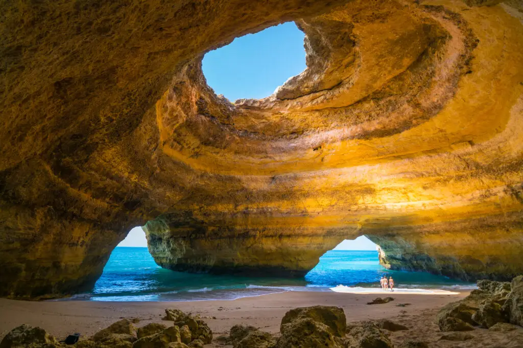 Tourists exploring the Benagil Sea Cave in Algarve, Portugal, with golden sandstone cliffs, turquoise ocean water, and sunlight streaming through the natural cave opening