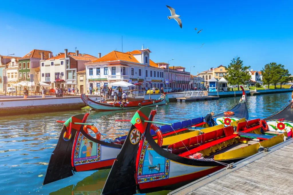 Colorful traditional Moliceiro boats on the canal in Aveiro, Portugal, with tourists exploring the historic waterfront and vibrant houses under a bright blue sky