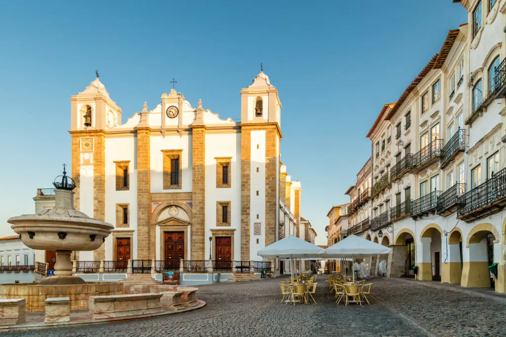 Giraldo Square and Antao Church in Evora, Portugal, showcasing a historic fountain, outdoor café seating, and traditional Portuguese architecture under a clear blue sky