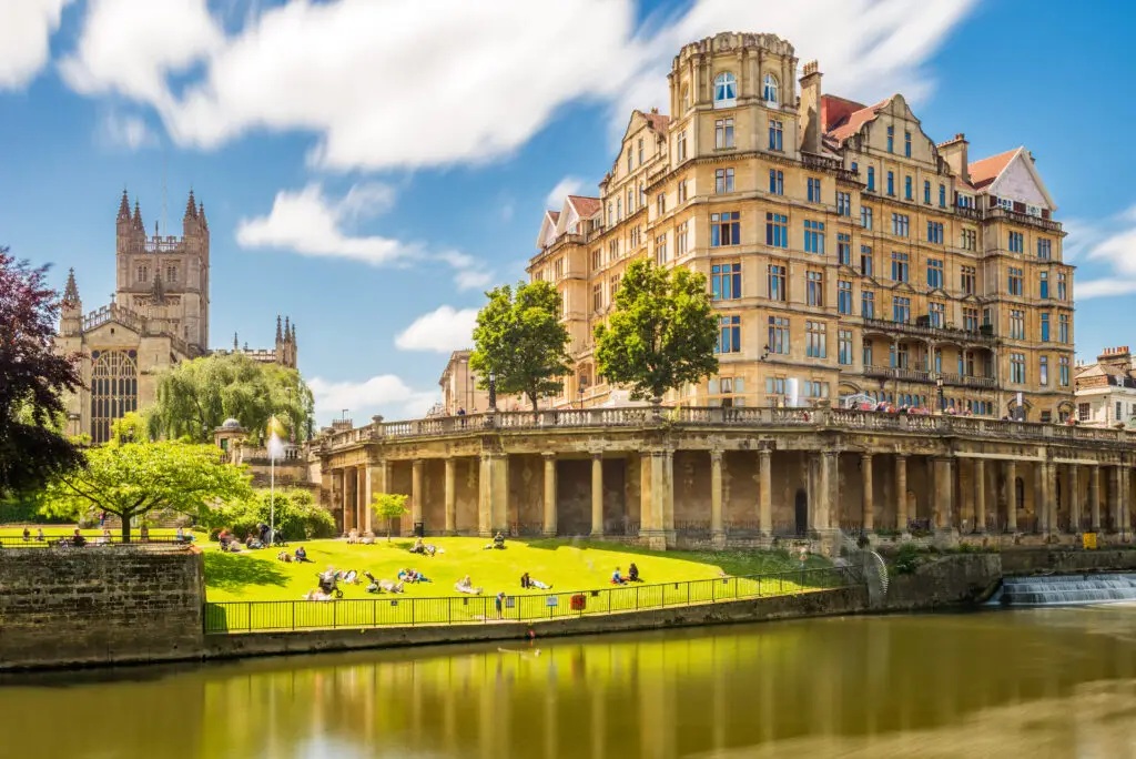 Pulteney Bridge and historic Palladian architecture along the River Avon in Bath, England, with Bath Abbey and tourists relaxing on the riverside on a sunny day