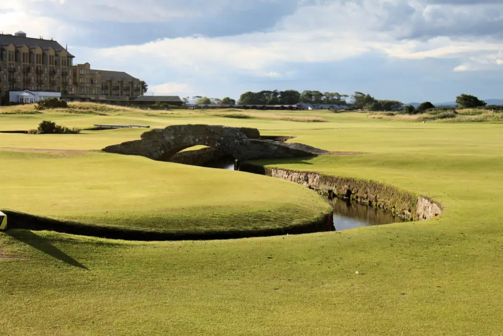 Stone bridge over the Swilcan Burn on the Old Course golf course in St Andrews, Scotland, with historic buildings in the background under a partly cloudy sky