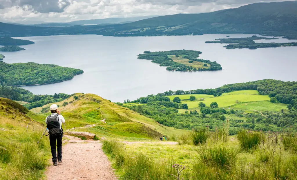 Hiker walking along the West Highland Way overlooking Loch Lomond in Scotland with lush green hills, scenic lake views, and islands in the distance