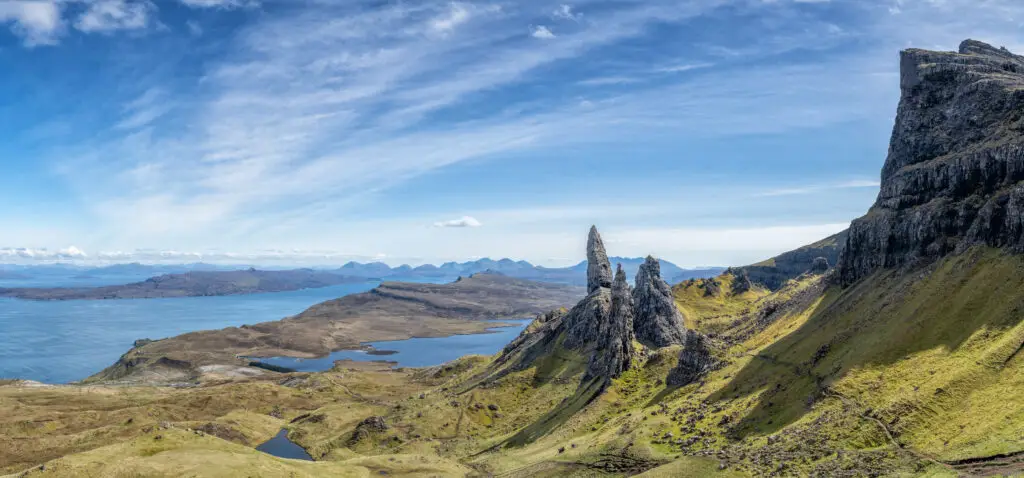Panoramic view of the Old Man of Storr rock formation on the Isle of Skye, Scotland, with rugged cliffs, rolling green hills, and blue coastal waters under a bright sky.