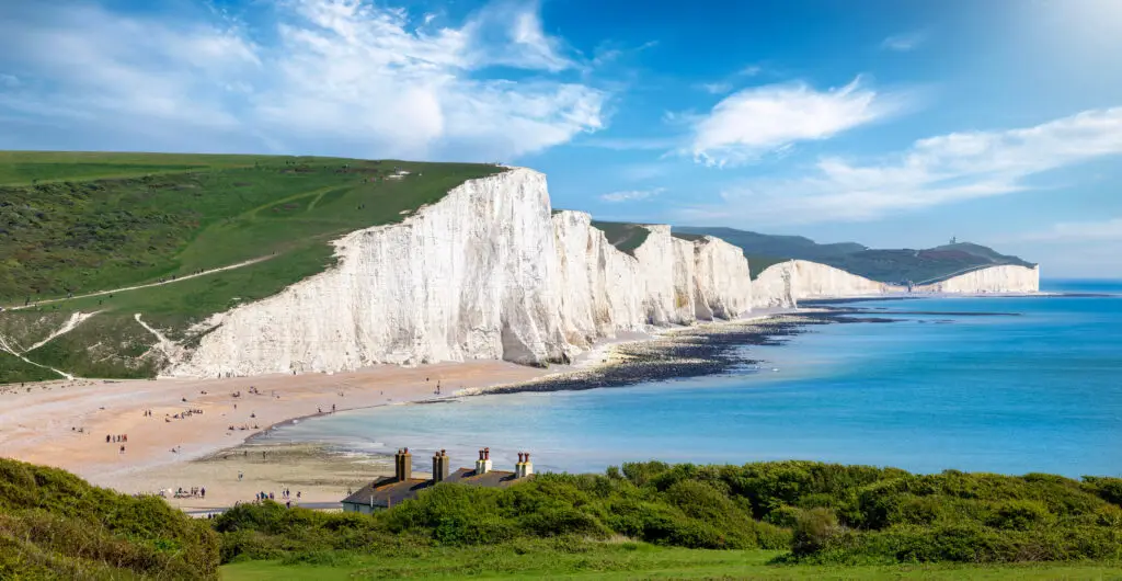 Panoramic view of the Seven Sisters Chalk Cliffs in Seaford, East Sussex, England, showing white cliffs, turquoise sea, green coastal hills, and tourists walking along the scenic beach and trails on a sunny summer day