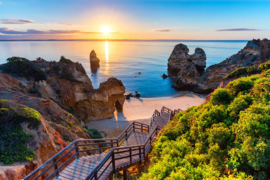 Wooden staircase leading down to Camilo Beach in Lagos, Algarve, Portugal with golden cliffs, turquoise Atlantic Ocean, and sunset view over the sea