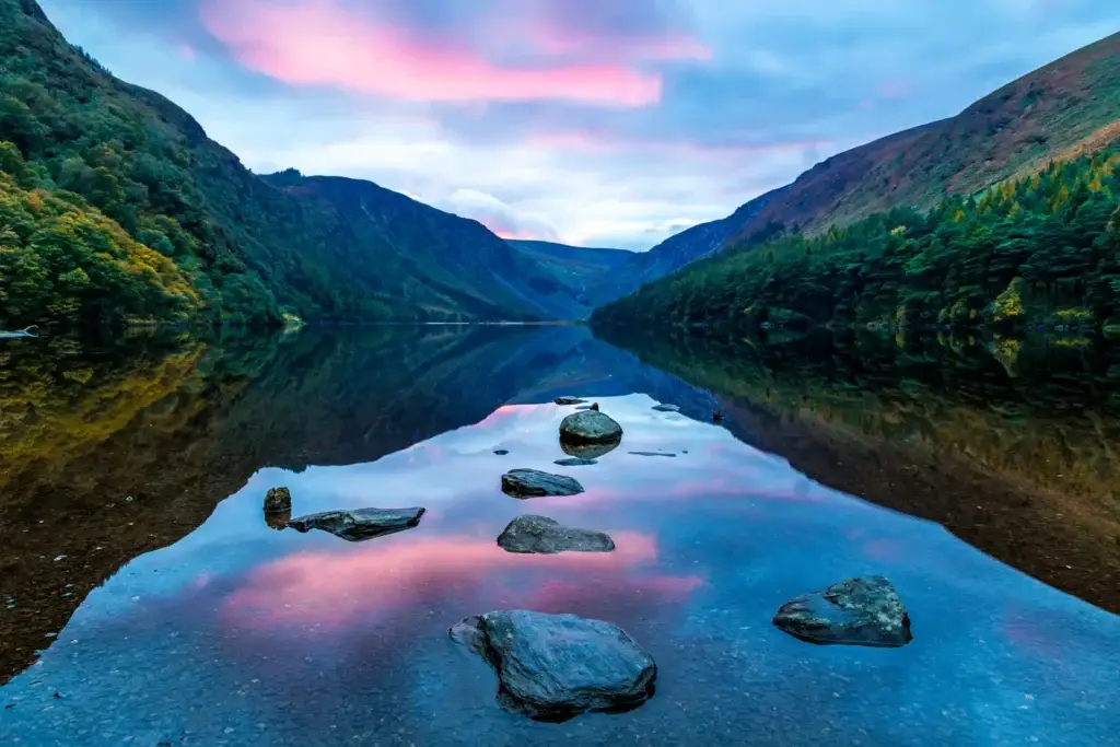 Scenic view of Glendalough Lake in County Wicklow, Ireland with calm reflective water, surrounding green mountains, and pink sunset sky