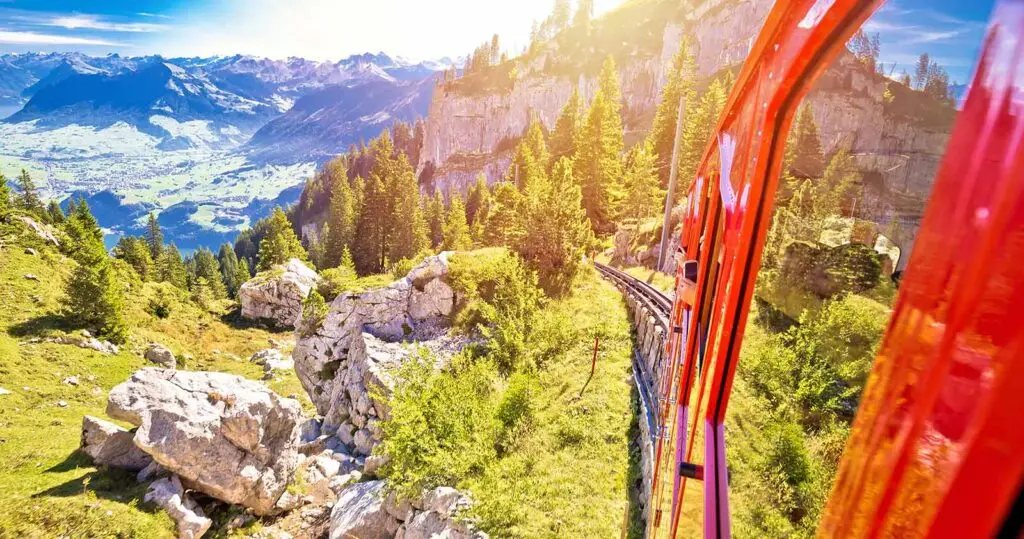 Scenic view from a red cogwheel train climbing Mount Pilatus in Switzerland with stunning alpine landscape, rocky cliffs, and green valleys under bright sunlight