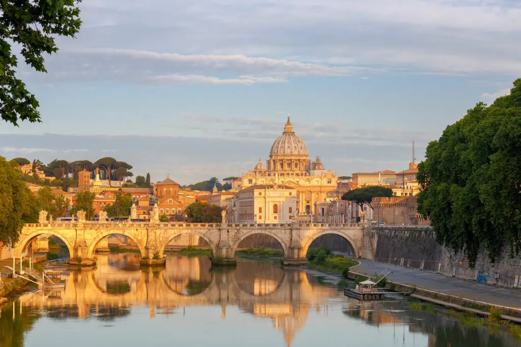 Saint Peter’s Cathedral in Vatican City, Italy, viewed from across the Tiber River with the historic bridge and cityscape at sunset