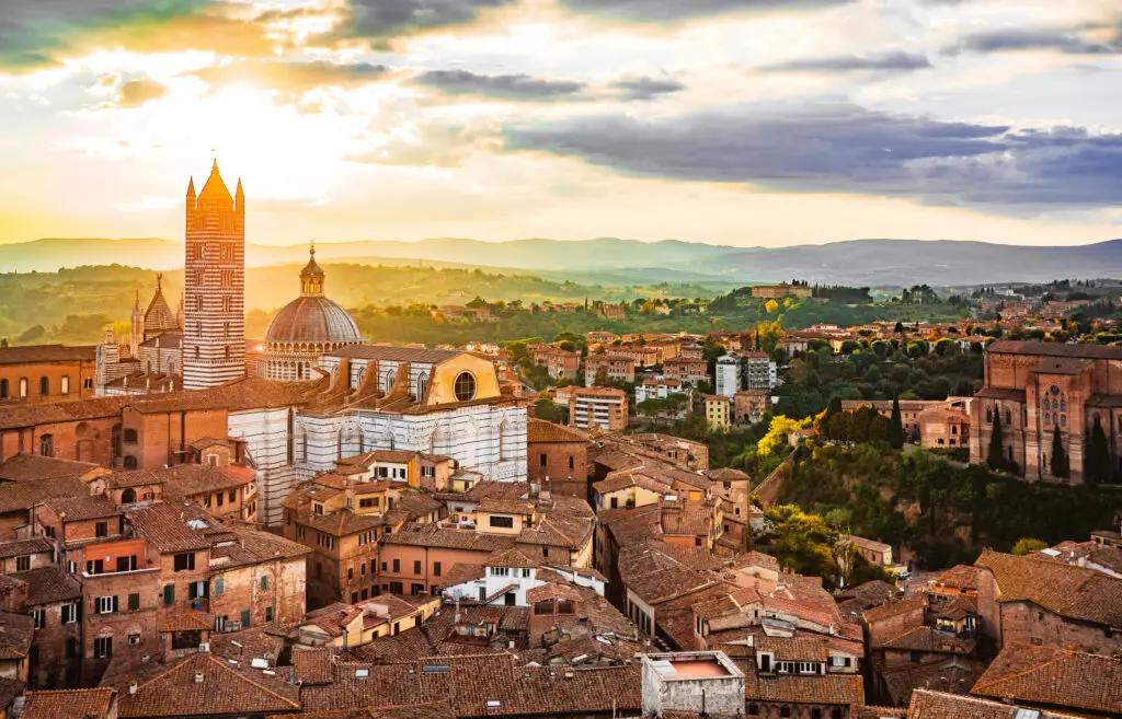 Panoramic view of Siena, Italy at sunset featuring the Siena Cathedral and historic city rooftops surrounded by Tuscan countryside.