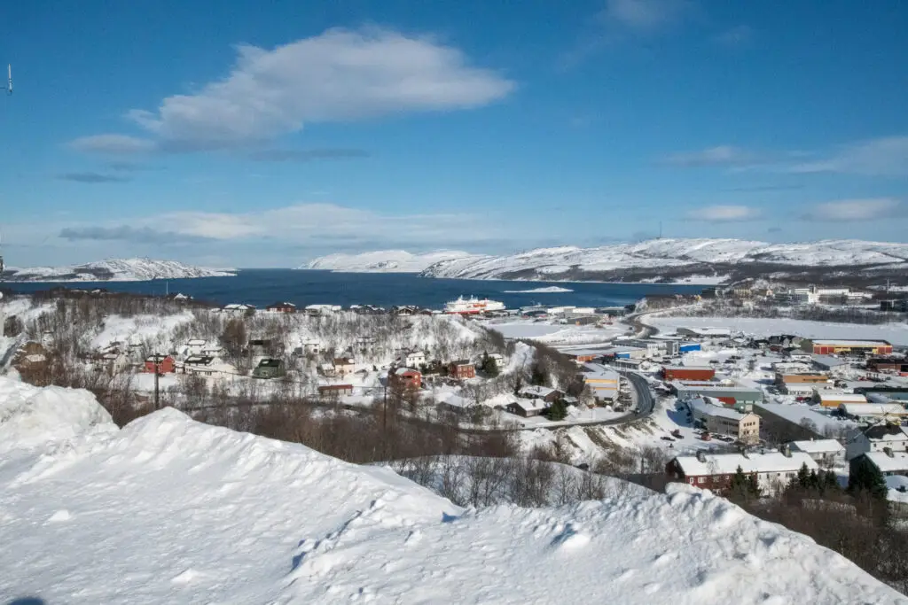 Winter landscape of Kirkenes, Norway with snow-covered houses, harbor, and fjord surrounded by Arctic mountains under a clear blue sky