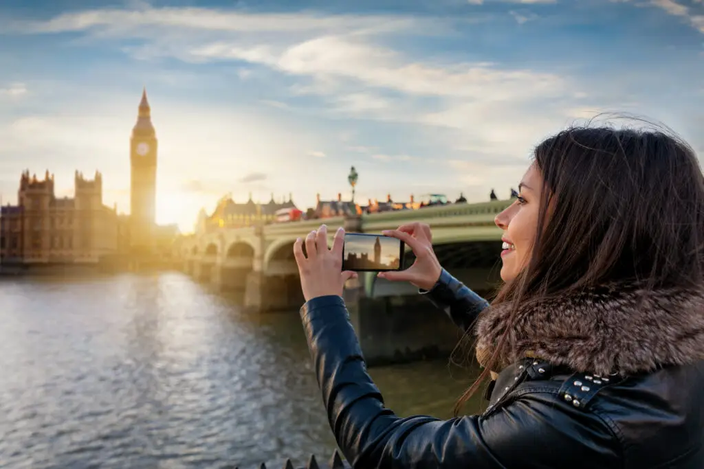 Young woman tourist taking a photo of Big Ben and Westminster Bridge in London at sunset during a European city tour