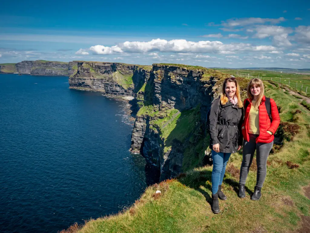 Two girls walking along the edge of the Cliffs of Moher in Ireland on a sunny day, enjoying scenic views of the Atlantic Ocean during a European private tour