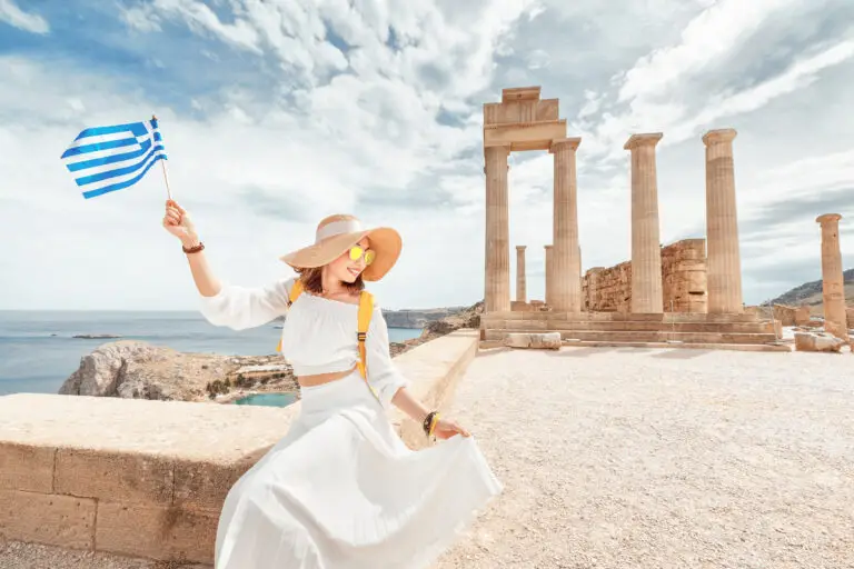 Woman holding a Greek flag at the ancient Acropolis of Lindos in Rhodes, Greece, with temple ruins and sea view in the background