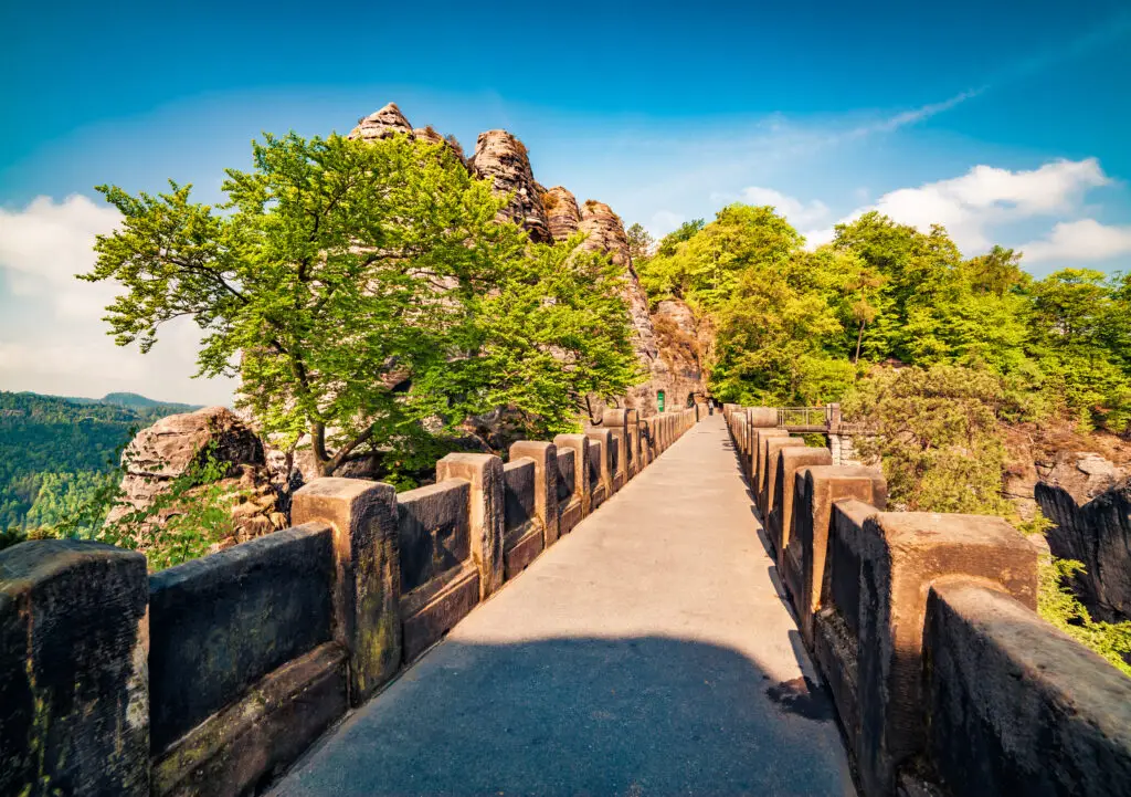 Scenic summer view of Bastei Bridge in Saxon Switzerland, Germany, surrounded by lush green trees and rocky cliffs under a bright blue sky