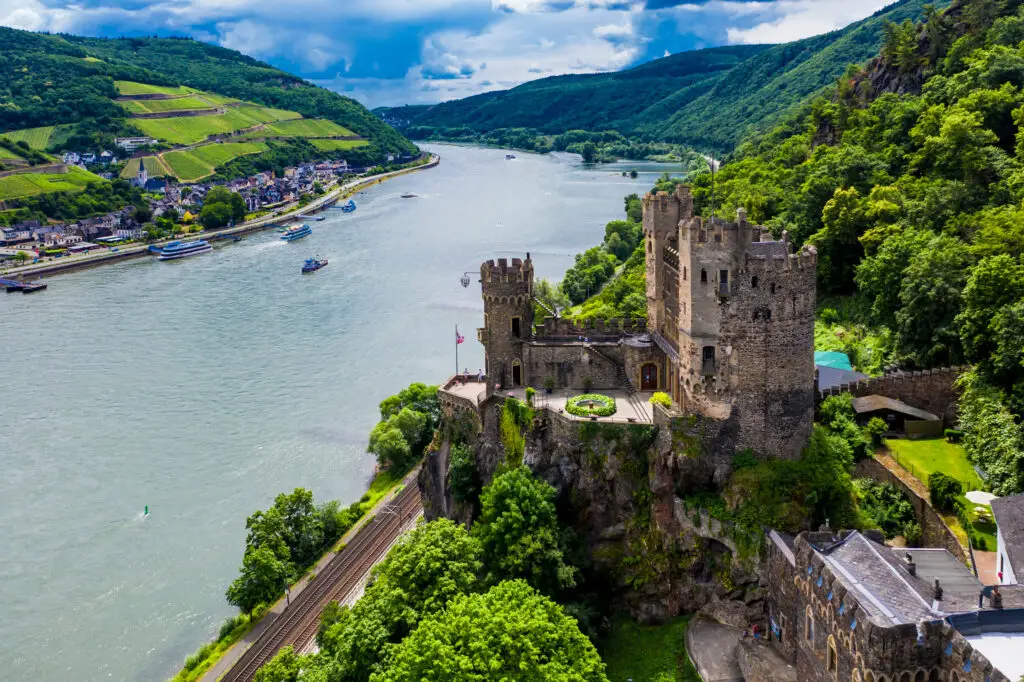 Rheinstein Castle overlooking the Rhine River in Germany with surrounding vineyards and riverboats, a popular European tourist attraction