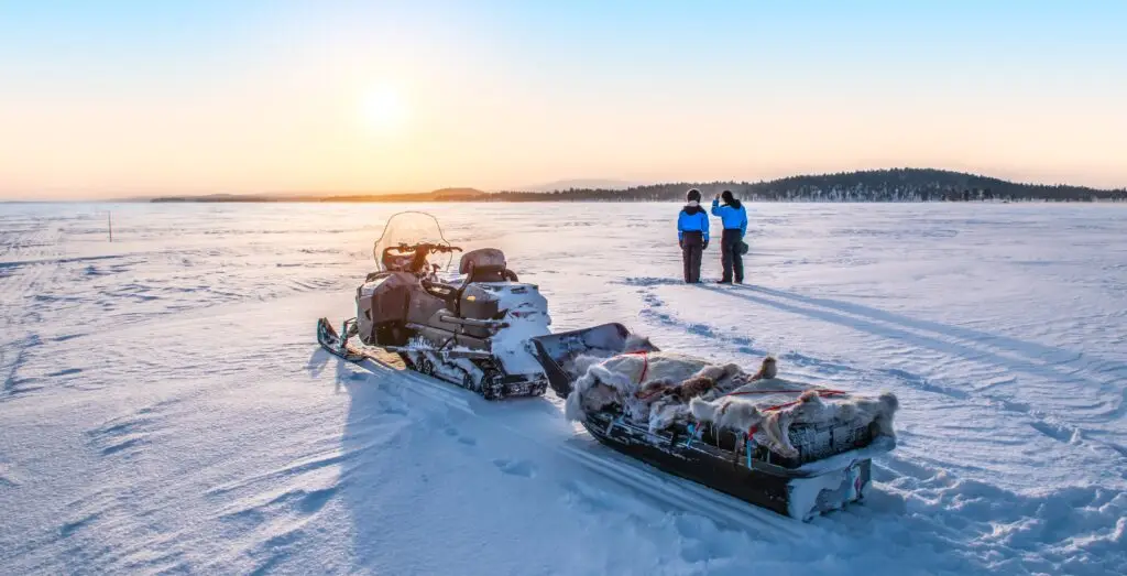 Young couple watching sunrise near snowmobile on frozen Lake Inari in Finnish Lapland during winter tour